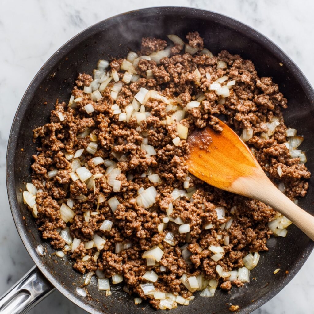 Ground beef and diced onions cooking in a skillet with a wooden spoon.