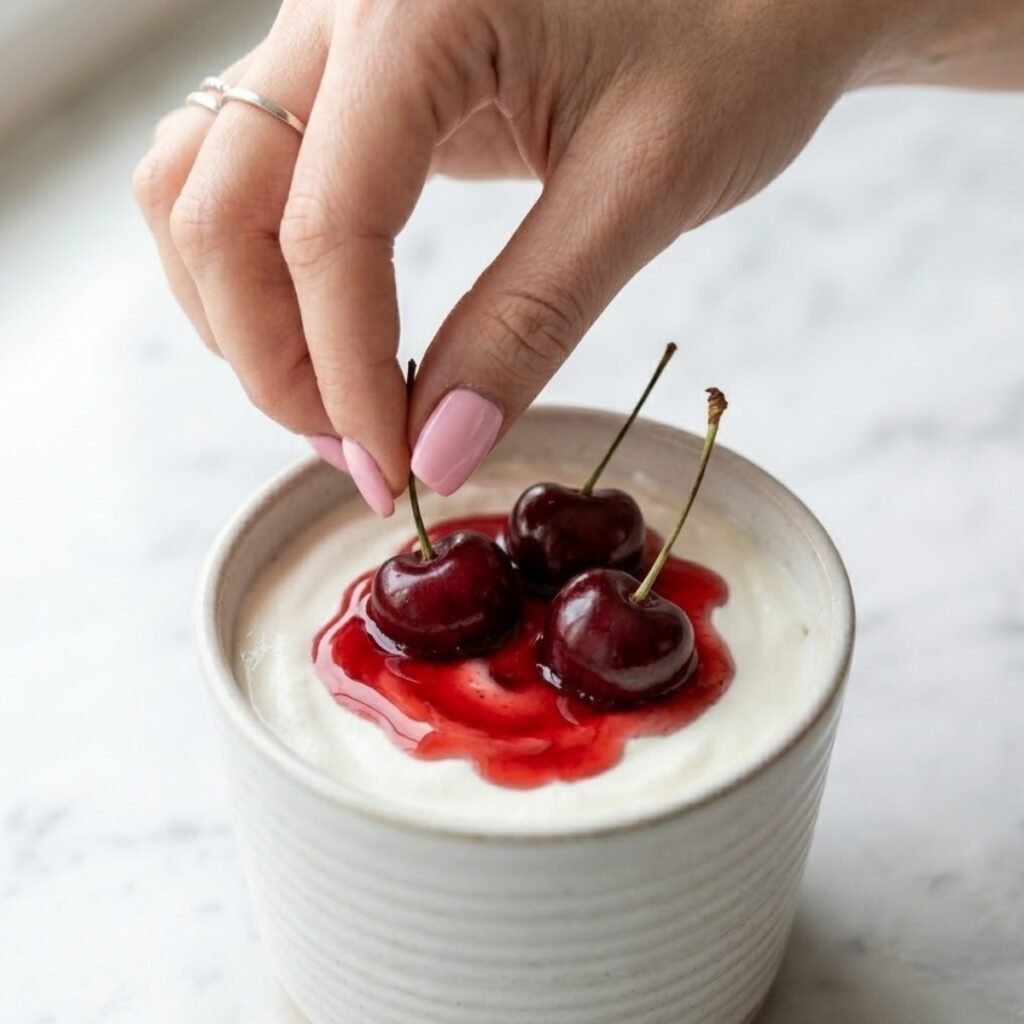 Hand with pink nails placing three fresh cherries on top of cherry syrup and whipped cream in a ribbed mug.