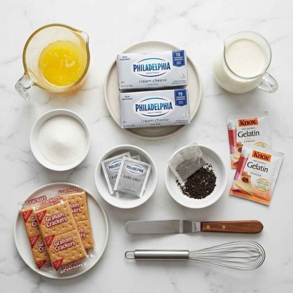 Square overhead view of raw ingredients for Earl Grey mousse cheesecake, including graham crackers, cream cheese, heavy cream, Earl Grey tea bags (steeping and opened), sugar, and gelatin powder, neatly arranged as separated components on a white marble counter. No hands.