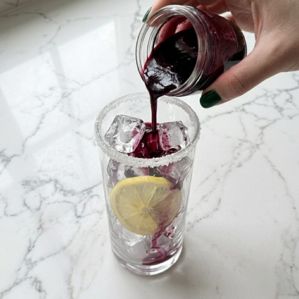 Deep purple blueberry syrup being poured over large ice cubes in a tall glass with a lemon wheel pressed against the side.