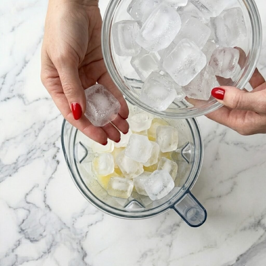 A top-down close-up view of a woman with classic red nails dropping frosty ice cubes from a large glass bowl into the blender canister filled with lemon liquid on a marble kitchen counter, creating a frozen lemonade slush.