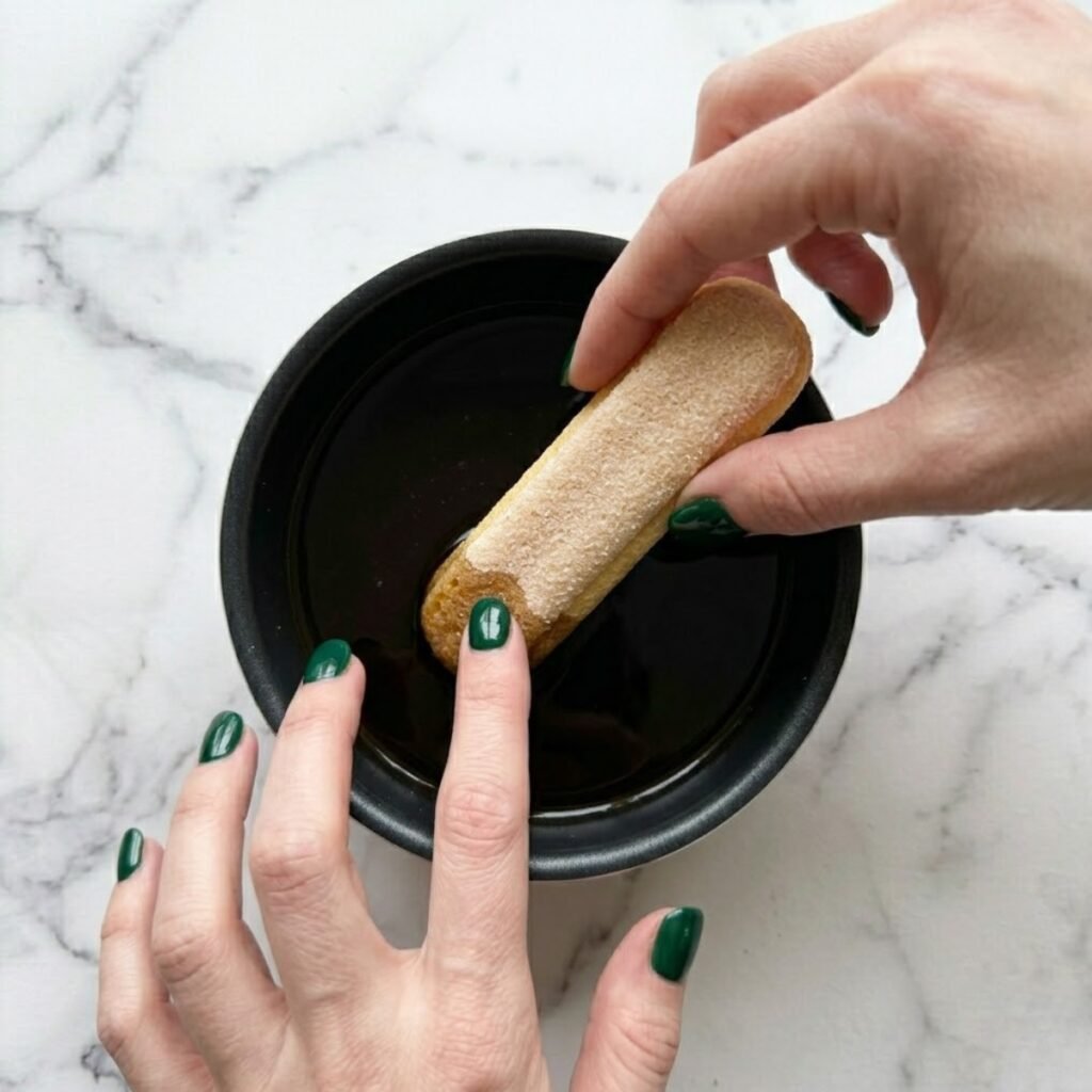 An overhead top-down close-up view of a woman with emerald green nails quickly dipping a ladyfinger biscuit into a shallow glass dish filled with dark, cooled espresso on a modern white marble counter.