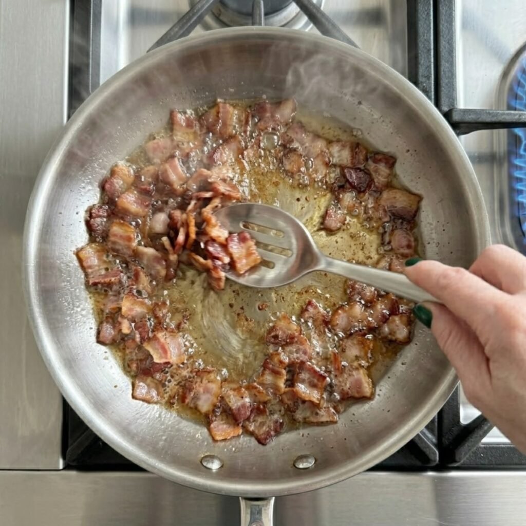 Sizzling bacon pieces being stirred in a wide stainless steel skillet on a gas stove with visible blue flames.