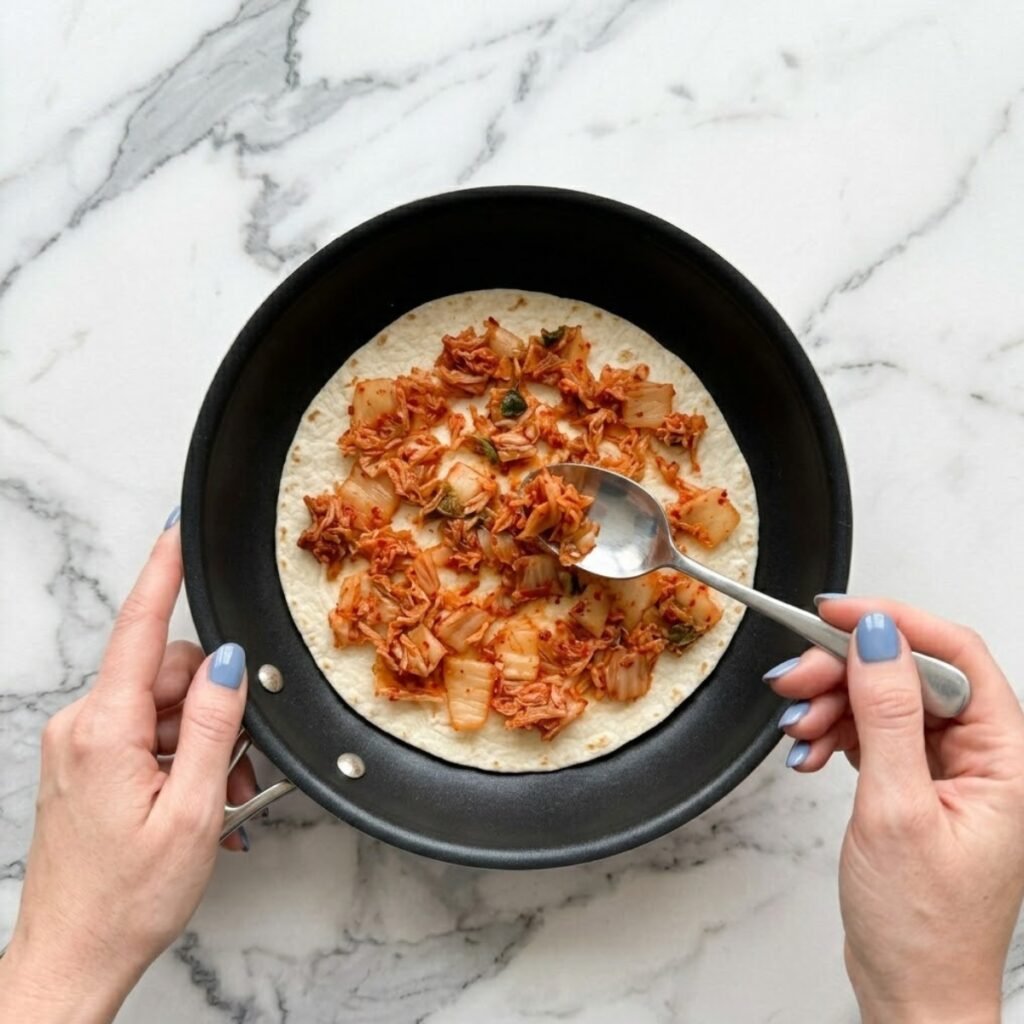 A close-up top-down view of a woman with Fog Blue nails placing a flour tortilla in a non-stick skillet and scattering finely chopped kimchi across it on a white marble kitchen counter.