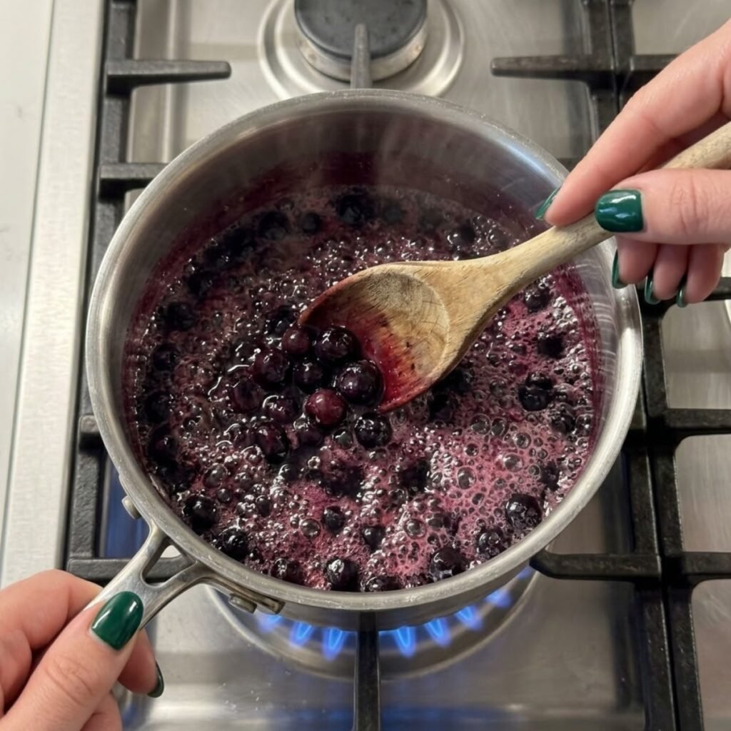 Close-up of fresh blueberries and sugar simmering in a small saucepan on a stovetop, with deep purple juice releasing into the liquid.