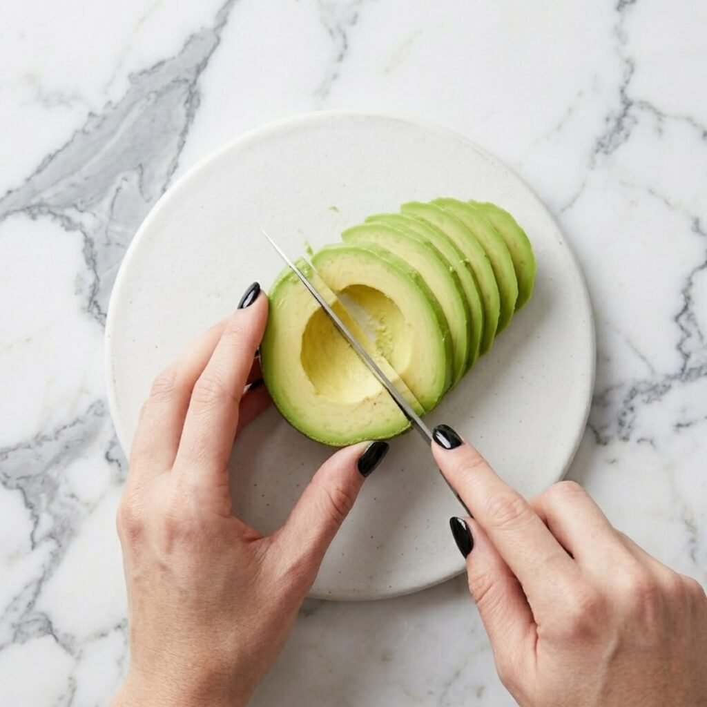 Close-up of hands with glossy black nails carefully slicing a ripe avocado half into even pieces on a marble surface.