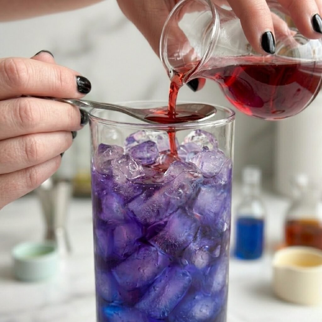 Close up of cranberry juice being poured over a bar spoon into a purple cocktail filled with crushed ice.