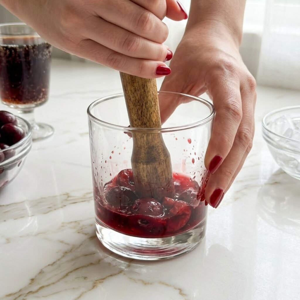 Instructional close-up showing the crucial step of muddling fresh cherries to extract juice and flavor for the cocktail base.