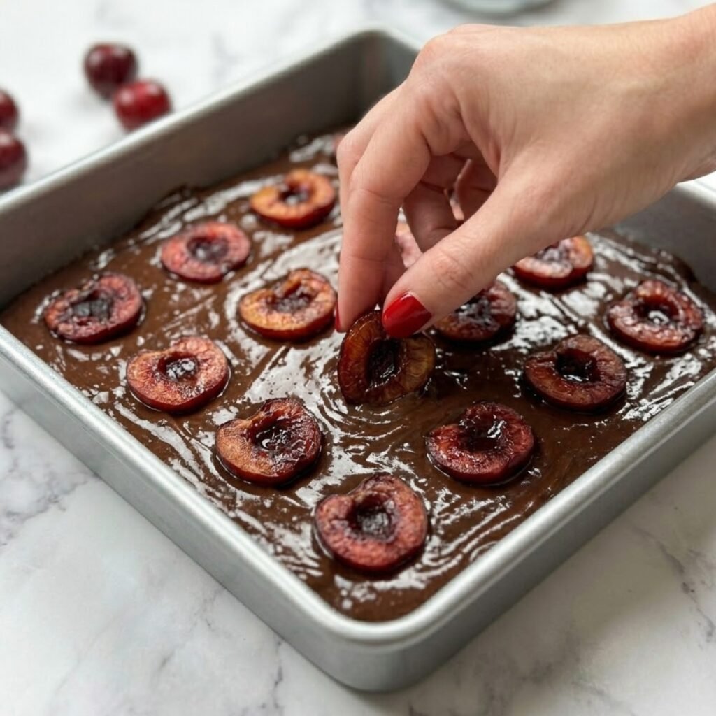 Close up of a hand with red nails gently pressing roasted cherry halves into thick dark chocolate brownie batter inside a square baking pan.
