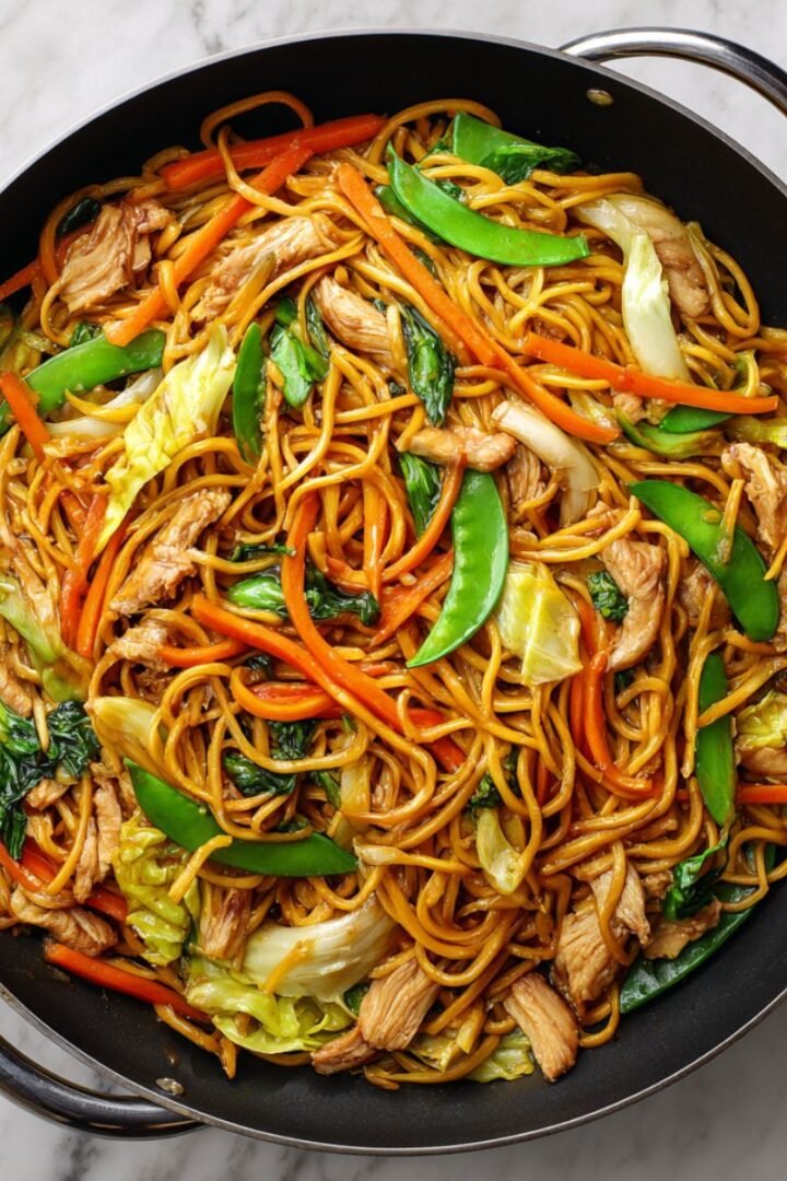 Chicken lo mein with soft golden noodles, tender chicken pieces and colorful vegetables in glossy soy sauce. Authentic Chinese stir fry with carrots, snow peas and cabbage in black wok.