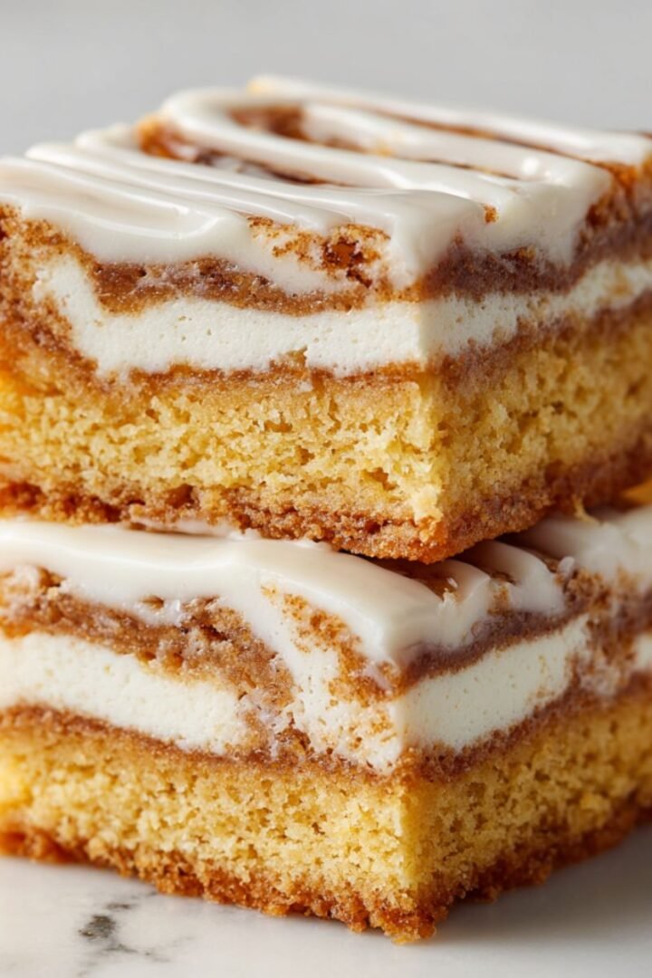 Cinnamon roll bliss bars with three layers - golden cake base, white cream cheese middle layer, and cinnamon cake top, all covered in thick vanilla glaze. Cut bars showing distinct cream cheese filling layer.