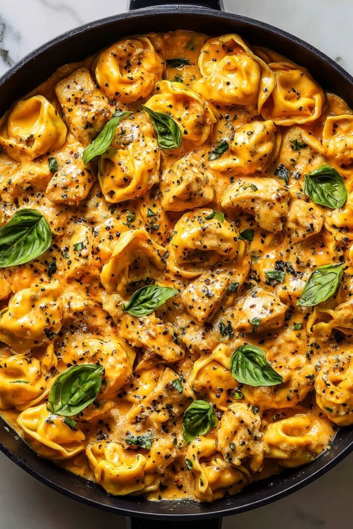 CrockPot chicken tortellini with cheese filled pasta, tender chicken pieces and creamy sauce garnished with fresh basil. Comfort food pasta dish in cast iron skillet photographed from above.
