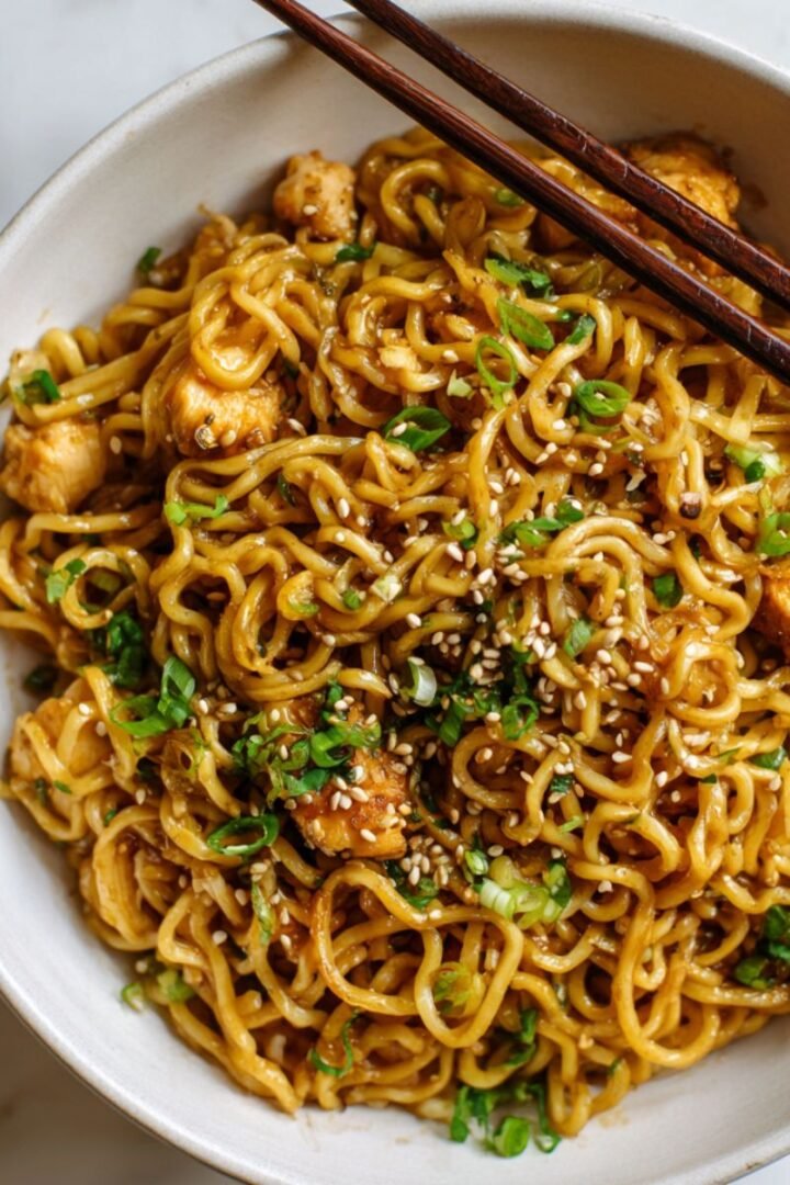 Garlic chicken noodles with glossy brown sauce and sesame seeds in white bowl. Savory 25 minute recipe with tender chicken pieces and chewy ramen noodles in sticky garlic sauce.