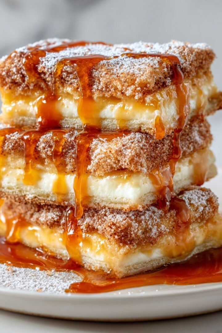 Three golden-brown churro cheesecake bars stacked on white plate showing flaky cinnamon-coated layers with creamy filling and amber caramel sauce drizzling down the sides