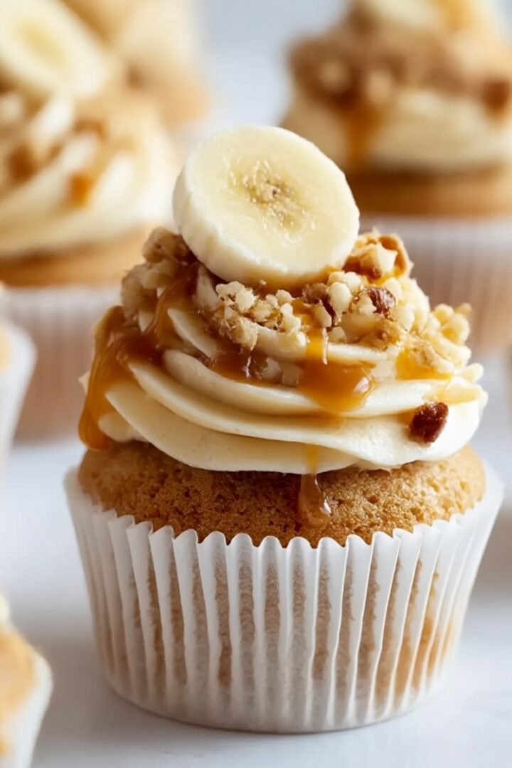 Banana pudding cupcake in white paper liner with moist banana cake, swirled vanilla pudding buttercream frosting, fresh banana slice on top, crushed vanilla wafer pieces, caramel drizzle, additional cupcakes softly blurred in background on white surface