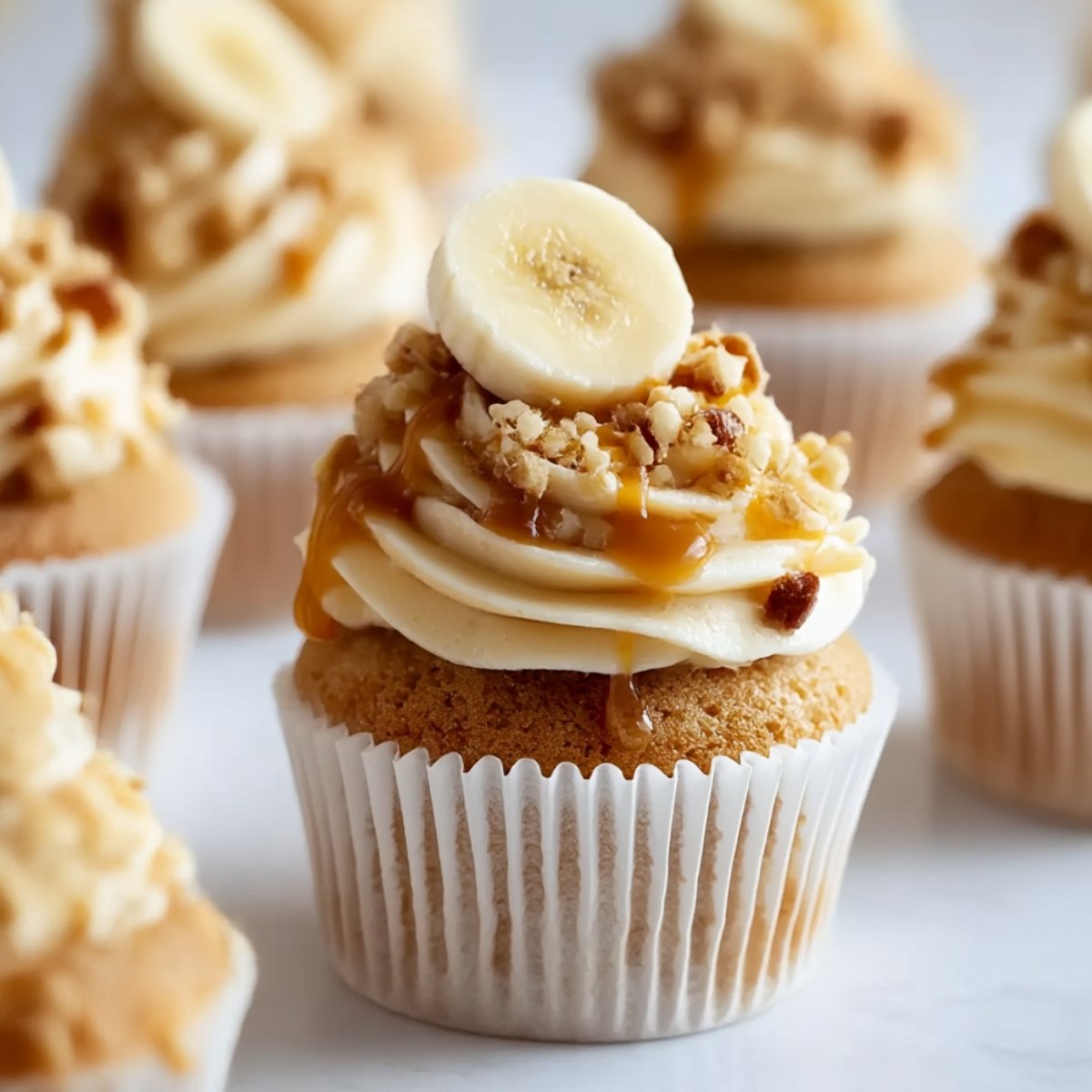 Banana pudding cupcake in white paper liner with moist banana cake, swirled vanilla pudding buttercream frosting, fresh banana slice on top, crushed vanilla wafer pieces, caramel drizzle, additional cupcakes softly blurred in background on white surface
