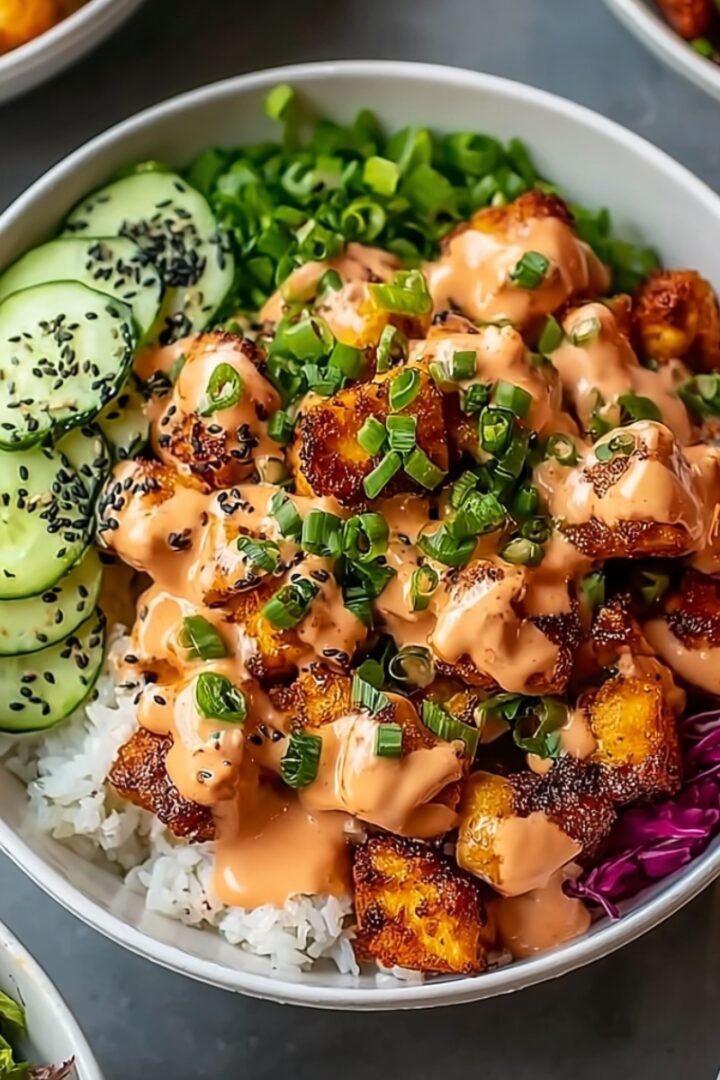 Bang Bang chicken bowl with white rice base, crispy golden chicken pieces drizzled with creamy spicy sauce, garnished with sliced green onions and black sesame seeds, accompanied by cucumber slices, edamame, and shredded purple cabbage