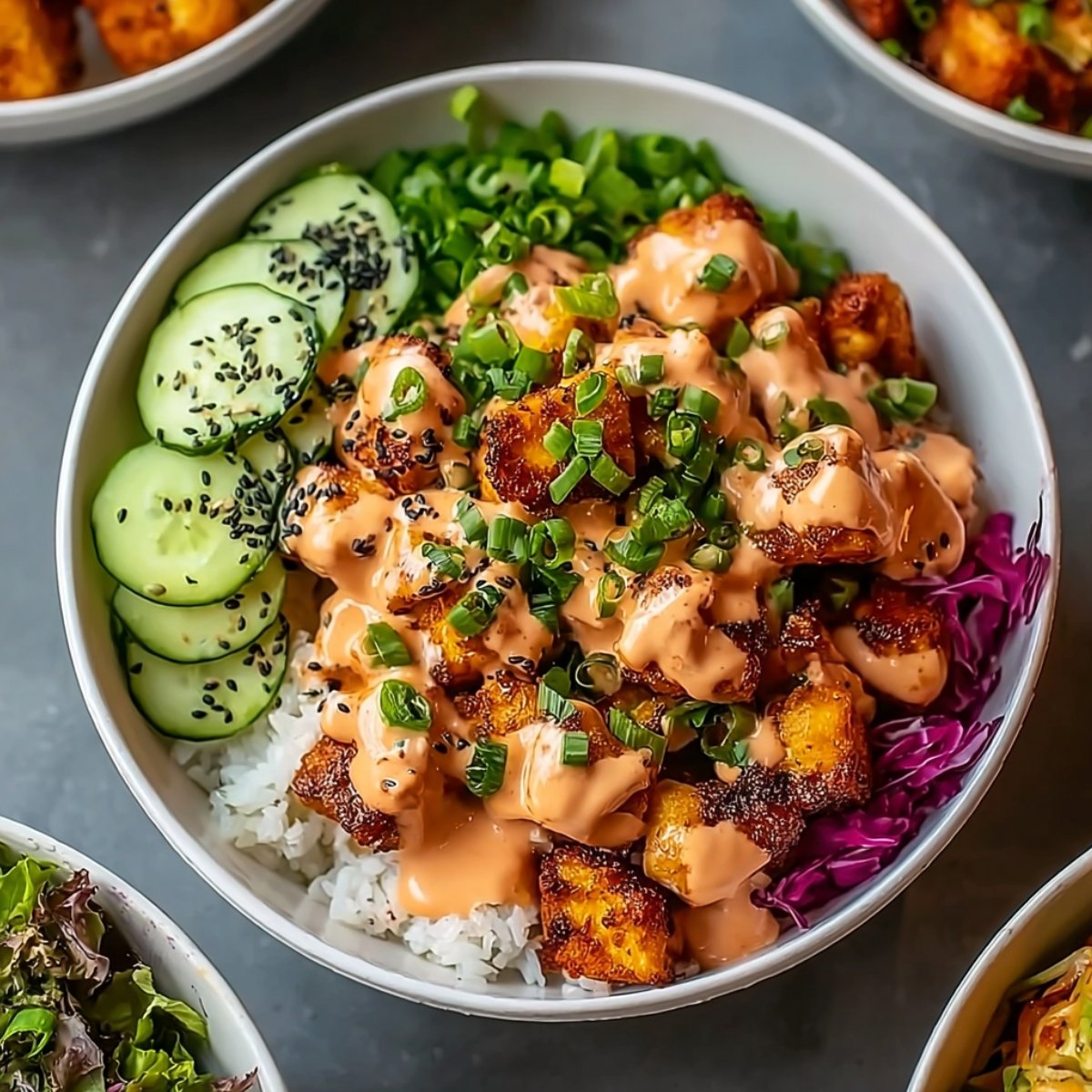 Bang Bang chicken bowl with white rice base, crispy golden chicken pieces drizzled with creamy spicy sauce, garnished with sliced green onions and black sesame seeds, accompanied by cucumber slices, edamame, and shredded purple cabbage