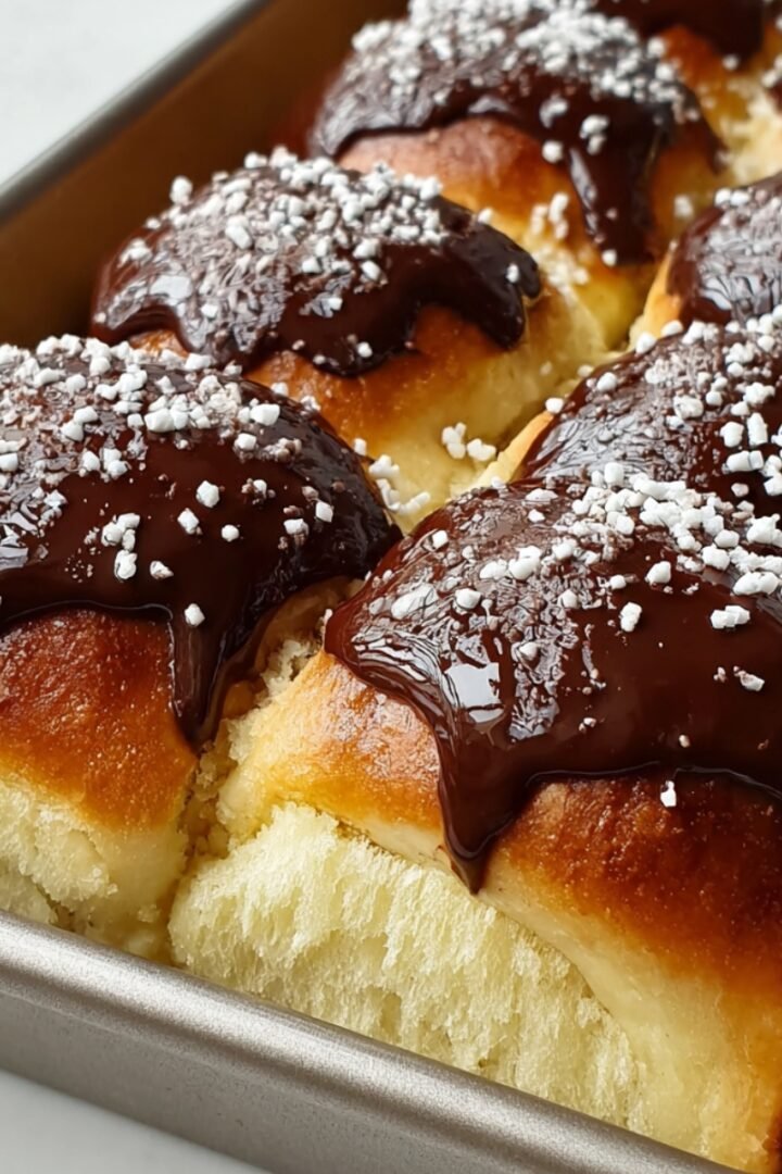 A metal baking pan filled with Boston Cream Pie Hawaiian rolls — soft golden sweet rolls sliced open to reveal a creamy vanilla custard filling, topped with glossy dark chocolate ganache and white sugar pearl sprinkles.