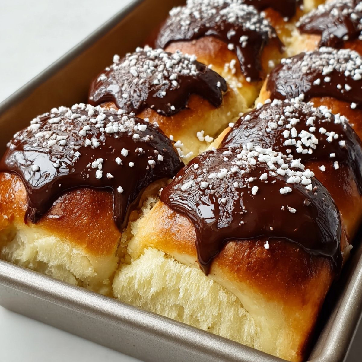A metal baking pan filled with Boston Cream Pie Hawaiian rolls — soft golden sweet rolls sliced open to reveal a creamy vanilla custard filling, topped with glossy dark chocolate ganache and white sugar pearl sprinkles.
