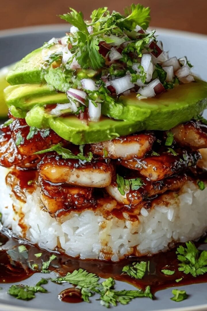 Honey lime chicken and avocado rice stack on blue-rimmed plate with white rice base, honey-glazed chicken pieces, sliced avocado, topped with diced red onion and fresh cilantro sprigs, drizzled with honey lime sauce, lime wedges on side, brown background