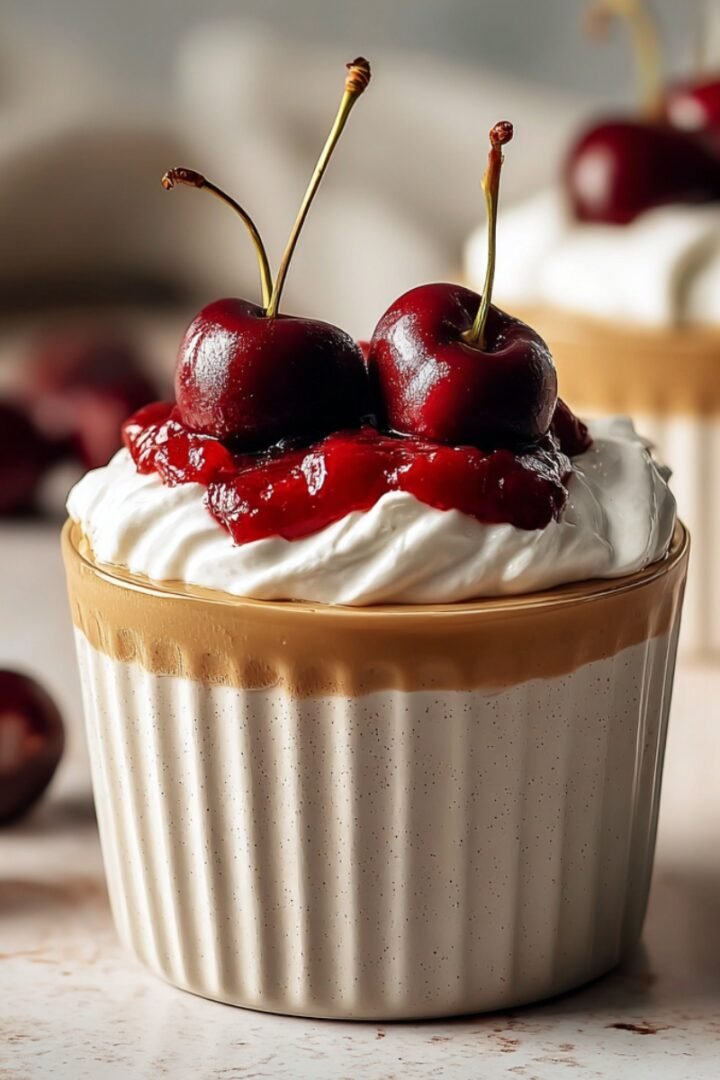 A ribbed ceramic cup of cherry vanilla latte topped with swirled whipped cream, cherry compote, and fresh whole cherries with stems, with loose cherries scattered around on a light surface.