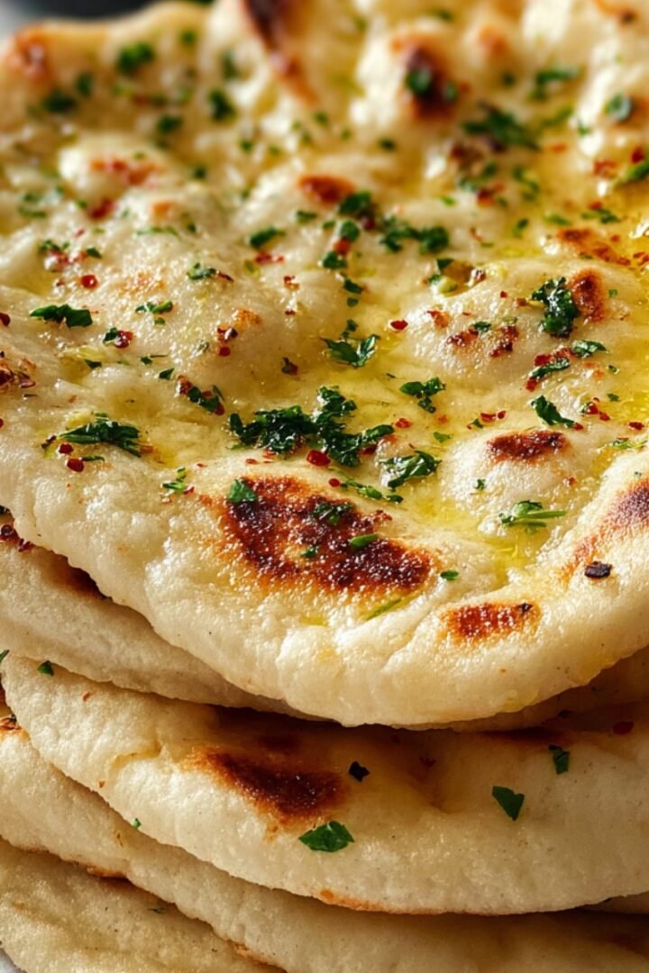 A stack of four soft Turkish flatbreads with golden char marks, the top piece brushed with melted butter and sprinkled with chopped fresh parsley and red pepper flakes, on a white surface.