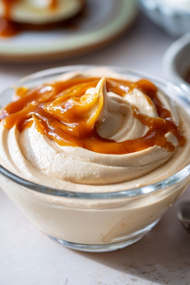 A glass bowl of swirled dulce de leche mousse drizzled with golden caramel sauce on top, with a spoon alongside and blurred serving dishes in the background on a light surface.