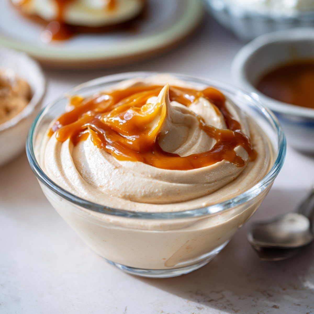 A glass bowl of swirled dulce de leche mousse drizzled with golden caramel sauce on top, with a spoon alongside and blurred serving dishes in the background on a light surface.