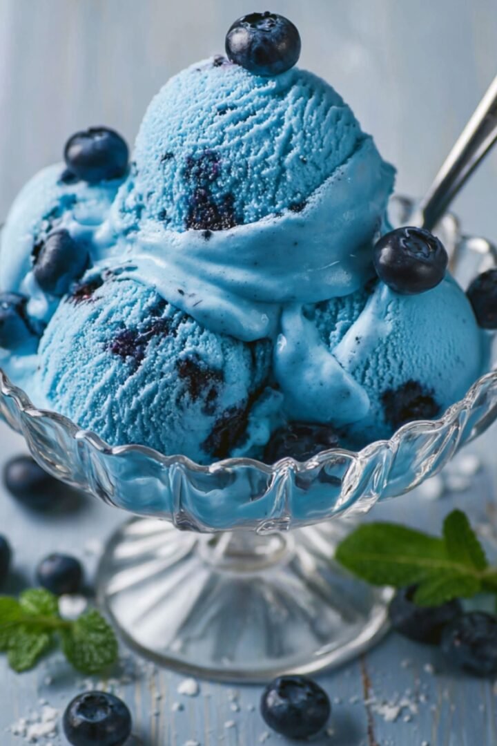 Scoops of vibrant blue ice cream with blueberry swirls served in a crystal glass dessert dish, topped with fresh blueberries and a spoon, with mint leaves and scattered blueberries on a light blue wooden surface.