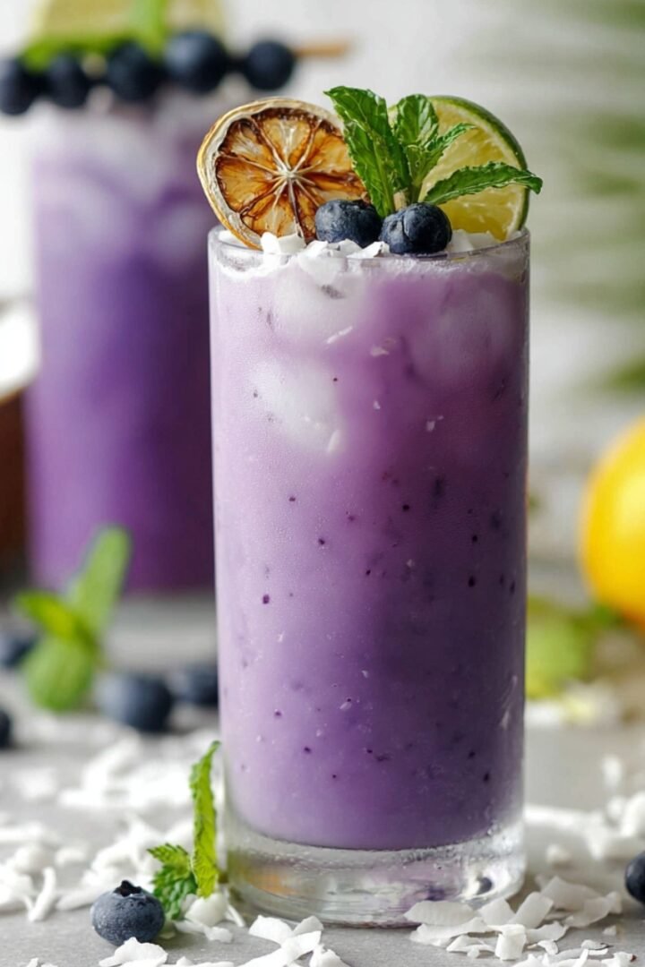 Two tall glasses of Blueberry Colada with a vibrant purple hue, topped with fresh blueberries, dried citrus slices, a lime wedge, and fresh mint, surrounded by shredded coconut.
