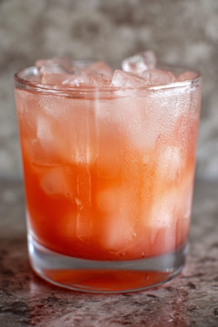A condensation-beaded rocks glass filled with a beautiful ombre guava white tea lemonade, graduating from a deep coral orange at the base to a soft blush pink at the top, packed with ice cubes, set on a speckled gray and rose granite surface with a softly blurred stone-textured background.