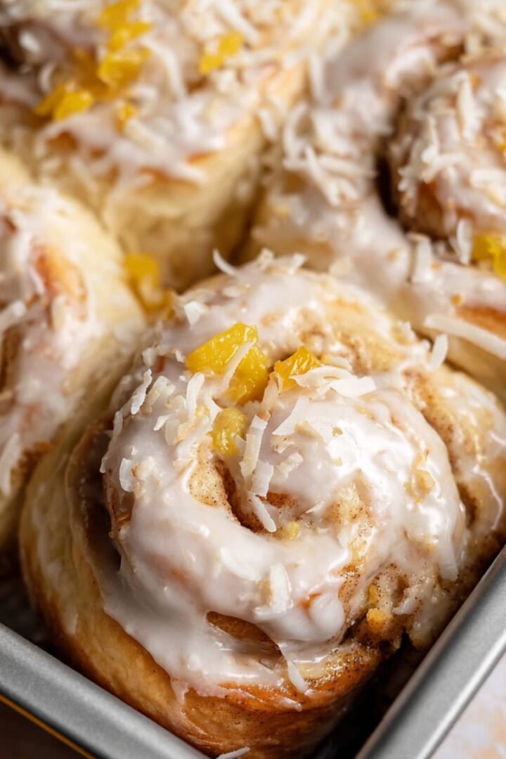 Fluffy baked cinnamon rolls in a metal pan, topped with creamy white glaze, shredded toasted coconut, and diced pineapple pieces.