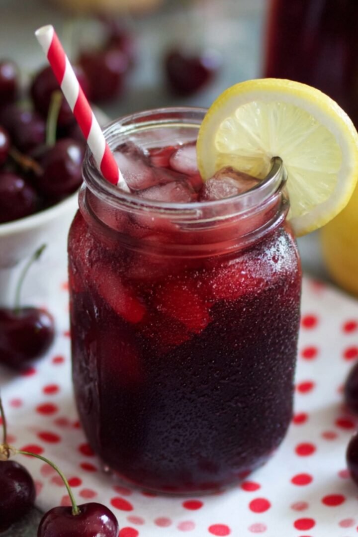 A mason jar of deep red Cherry Iced Tea over ice, garnished with a lemon slice and a red striped straw, surrounded by fresh cherries and lemons.