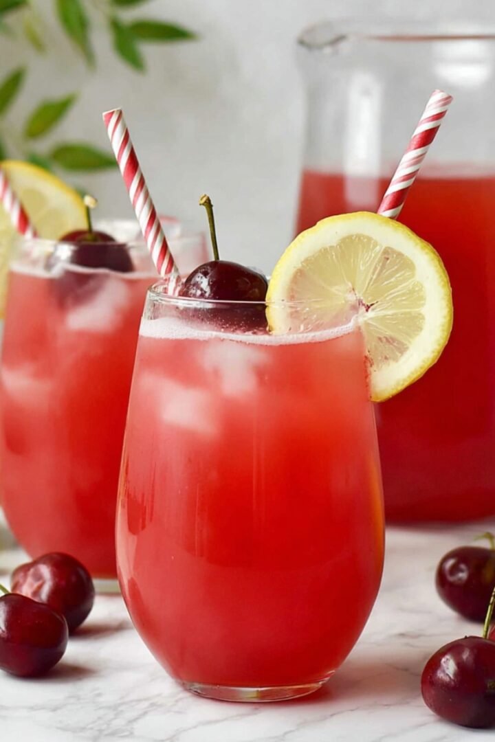 Two glasses of vibrant red cherry lemonade over ice, garnished with fresh cherries, lemon slices, and red and white striped paper straws, with a pitcher of cherry lemonade, whole lemons, and scattered cherries on a marble surface.