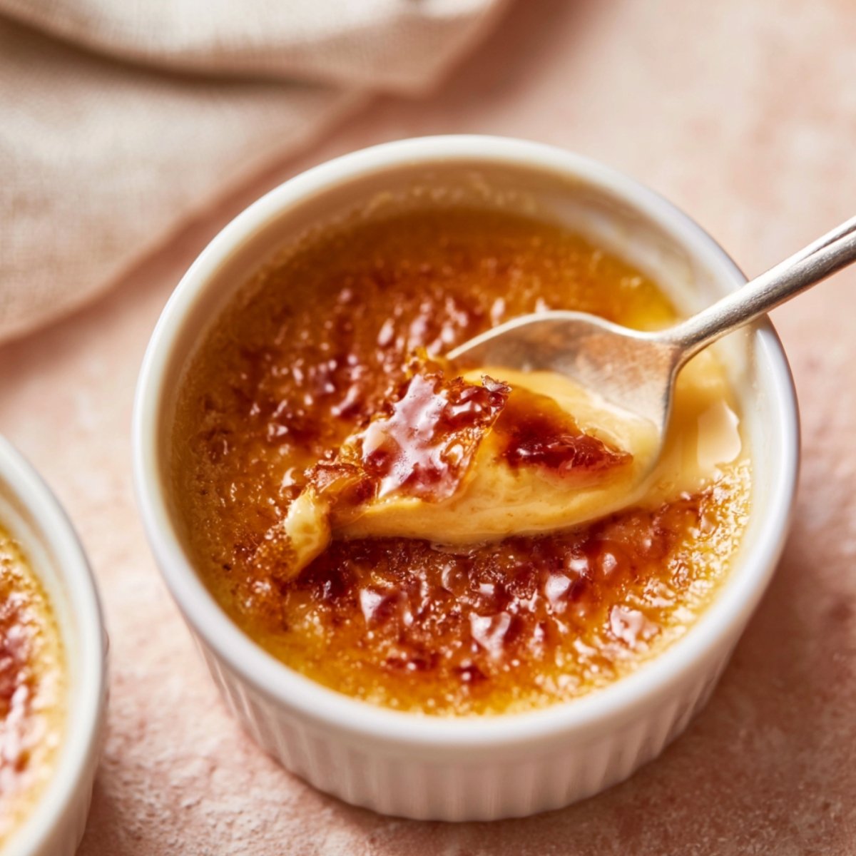 A white ramekin of coffee crème brûlée with a cracked caramelized sugar crust being broken by a spoon to reveal the creamy custard beneath, with a second ramekin visible alongside on a pink surface.